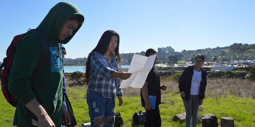 A group of four high school students stand in a line outdoors while leading a presentation about the natural environment 