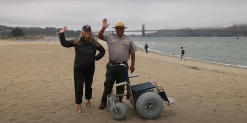 A Parks Conservancy staff member and a National Park Service staff member stand waving and smiling on East Beach next to a beach wheelchair.