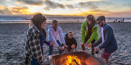Ocean Beach visitors warm up beside a bonfire in the iconic fire pits as a golden sunset illuminates the sky.