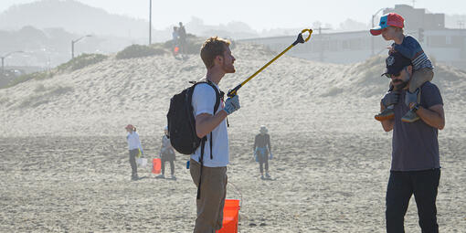 Adult shows trash to curious boy on his dad's shoulders on Ocean Beach in San Francisco.