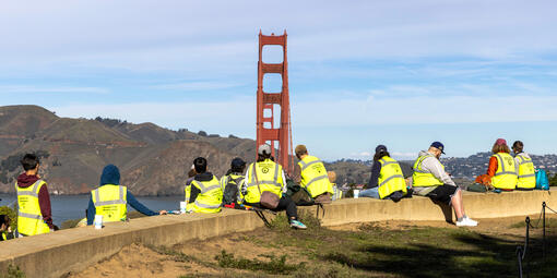 A group of volunteers in bright vests sit on a presidio bench with the golden gate bridge and marin headlands in view.
