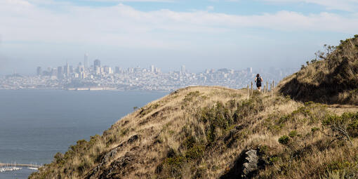 A park visitor runs on the SCA Trail in the Marin Headlands with the San Francisco skyline in background.