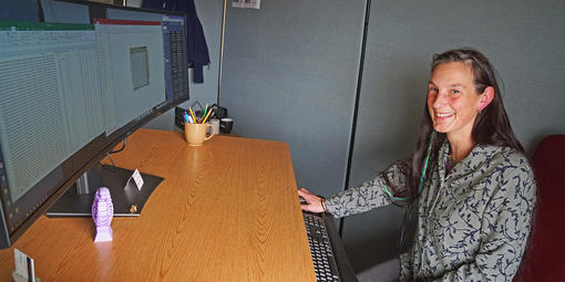 Lizzy Edson smiles behind a large curved computer monitor.