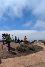Volunteers in orange vests look through scopes and binoculars on a brown hilltop under partiall cloudy sky.