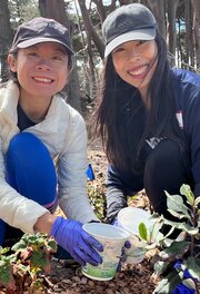 Two volunteers kneel and smile for the camera while collecting weeds.